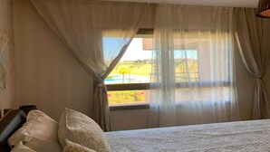 Een slaapkamer met een bed, gordijnen en een raam met uitzicht op een zwembad en groen.