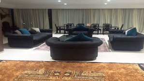 Amplia sala de estar con sofás oscuros y una mesa de centro, un comedor con mesa y sillas, y un gran ventanal con cortinas.