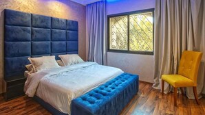A bedroom with a large bed, a tufted headboard, a window with decorative iron bars, a yellow chair, and a wooden floor. - High standing villa with swimming pool 5231 (Marrakech)