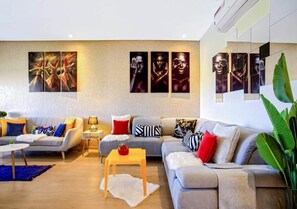 A modern living room with a grey sofa, a small wooden table, and a coffee table. There are multiple cushions and a plant on the sofa. - Splendid apartment in Prestigia Marrakech 5234 (Marrakech)