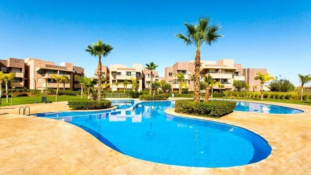 A swimming pool with a curved design, surrounded by palm trees and a building complex in the background. - Splendid apartment in Prestigia Marrakech 5234 (Marrakech)