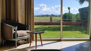Una habitación con un ventanal grande, un sofá, una mesita y vistas a un espacio verde con árboles y un edificio a lo lejos.
