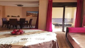 A hotel room with a bed, a dining table with chairs, a balcony with red curtains, and a painting on the wall. - Pleasant apartment in Premium Village Marrakech 5239 (Marrakech)