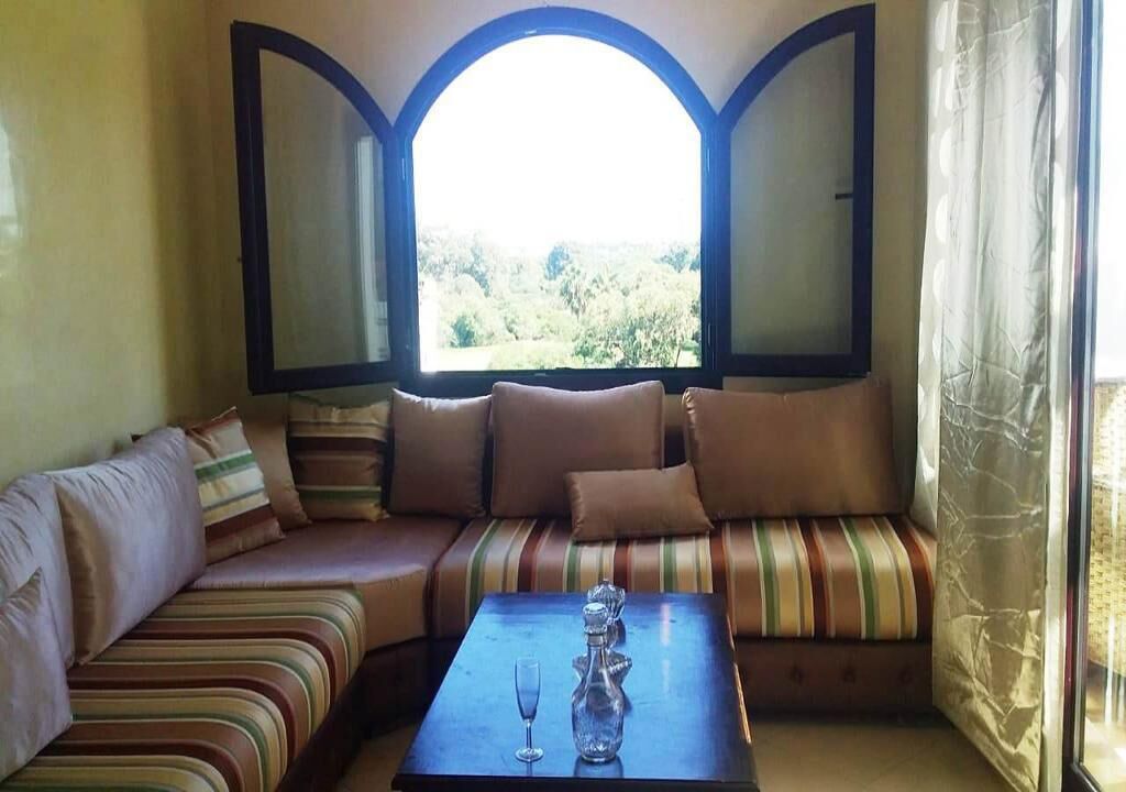 A cozy seating area with a cushioned bench and a small table, a large arched window with curtains, and a view of greenery outside.