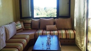 Un coin salon confortable avec un banc rembourré et une petite table, une grande fenêtre en arc surmontée de rideaux et une vue sur la verdure à l’extérieur.