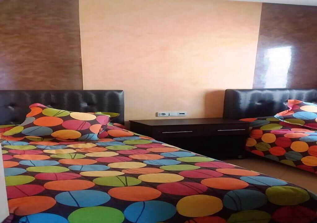Ein Hotelzimmer mit zwei Betten, jeweils mit einer farbenfrohen, kreisenden Musterdecke. Dazwischen befindet sich ein kleiner Tisch mit einer Steckdose und einem kleinen Gerät.
