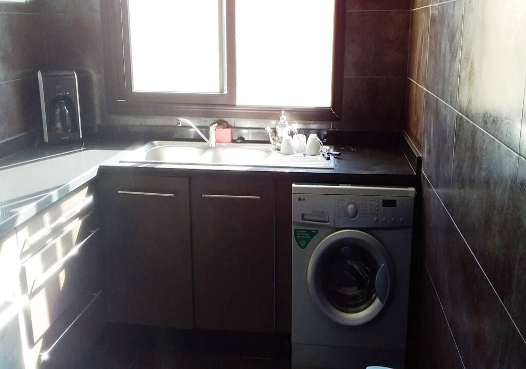 A compact kitchen with a built-in washer and dryer, a window, and a countertop with a kettle and cups.