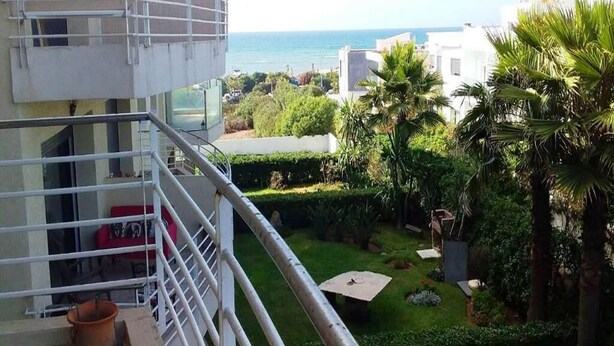 Un balcón con vista al mar, un pequeño jardín con palmeras y un edificio blanco al fondo.
