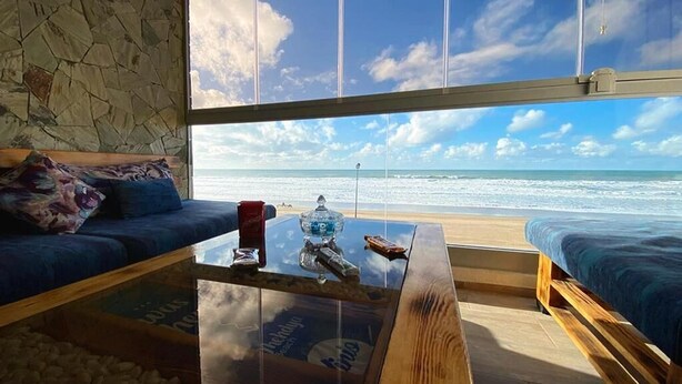 Une pièce dotée d’une grande fenêtre donnant sur une plage, une table basse en verre sur laquelle sont posés divers objets, et un canapé agrémenté de coussins.