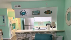 Una sala de estar con un sillón verde azulado, una cocina con un refrigerador, y un comedor con taburetes de bar.