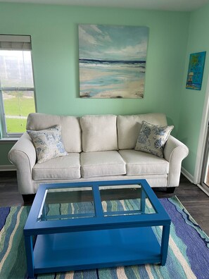 Una sala de estar con un sofá color beige, una mesa de centro azul y un cuadro de una playa.