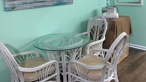 Un comedor con una mesa de vidrio rodeada de sillas de mimbre blanco, un cuadro de un velero en la pared y un espejo sobre la mesa.