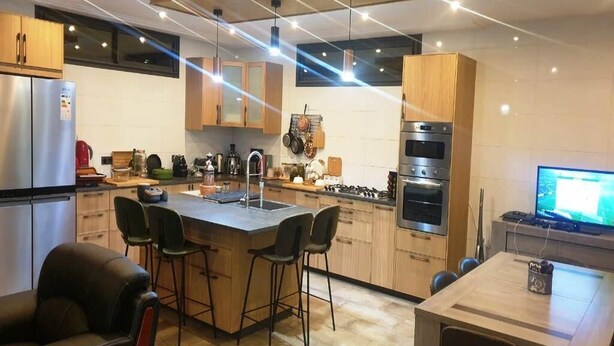 Una cocina moderna con una isla central, taburetes de bar, una mesa de comedor y un televisor.