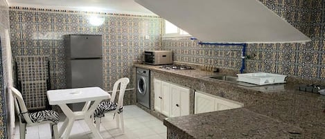 A compact kitchen with patterned tiles, a refrigerator, a microwave, a washing machine, and a dining area with chairs.