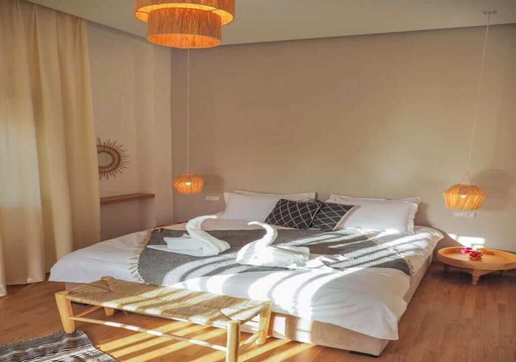 Un llit ben fet amb un coixí amb forma de cigne, una banca, una tauleta de nit amb una làmpada i una decoració de sol a la paret.