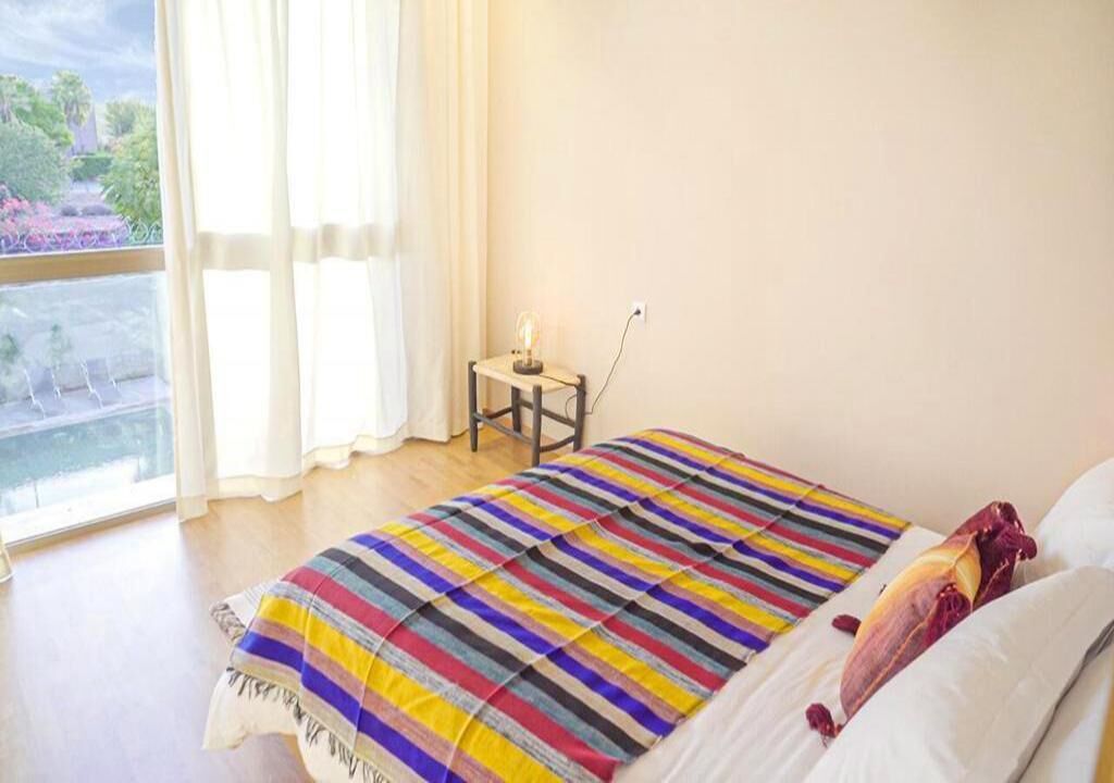 A bedroom with a bed, a small table with a lamp, and a view of the outdoors through a window.