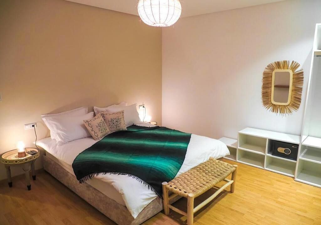 Une chambre à coucher comprenant un lit, des lampes de chevet, une table de chevet, un banc en osier et un miroir décoratif accroché au mur.