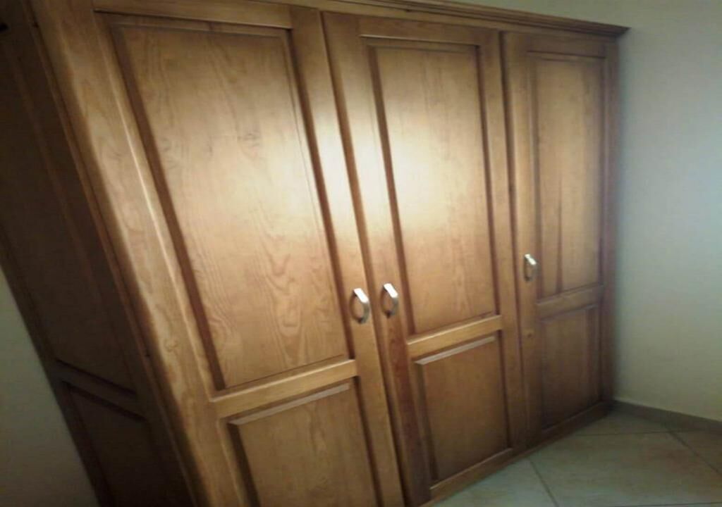 Un armario de madera con dos puertas y una repisa, situado en un rincón de una habitación con suelo de baldosas.