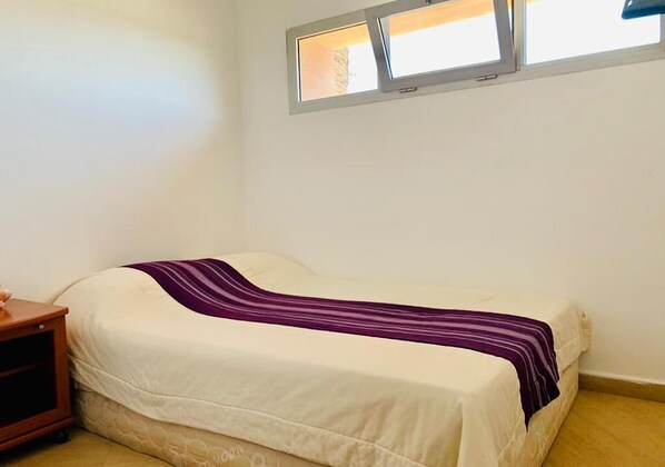 Un lit simple avec une couverture rayée blanche et violette, une table de chevet en bois et une fenêtre donnant sur l’extérieur.