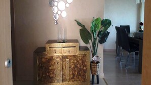A modern living room with a decorative wall piece, a wooden cabinet with drawers, a potted plant, and a dining area in the background. - Superb apartment in Prestigia Marrakech 5315 (Marrakech)