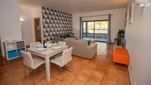 A modern living room with a sofa, dining table, and chairs. There is a window with a view of trees and a balcony. - Corbo apartment (Lagos)