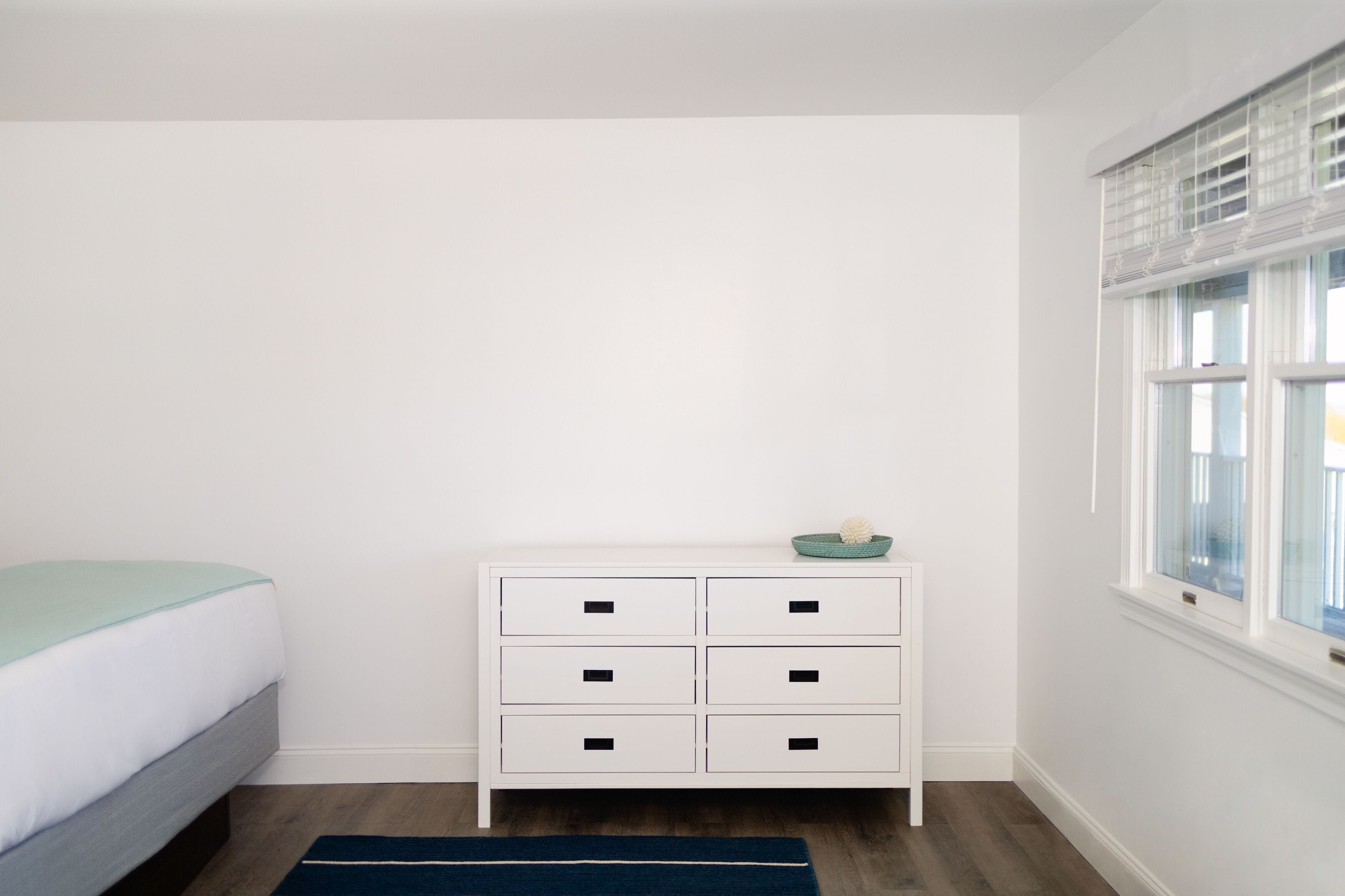 Quarto com cama, cômoda branca com gavetas, tapete azul escuro e janela com persianas.