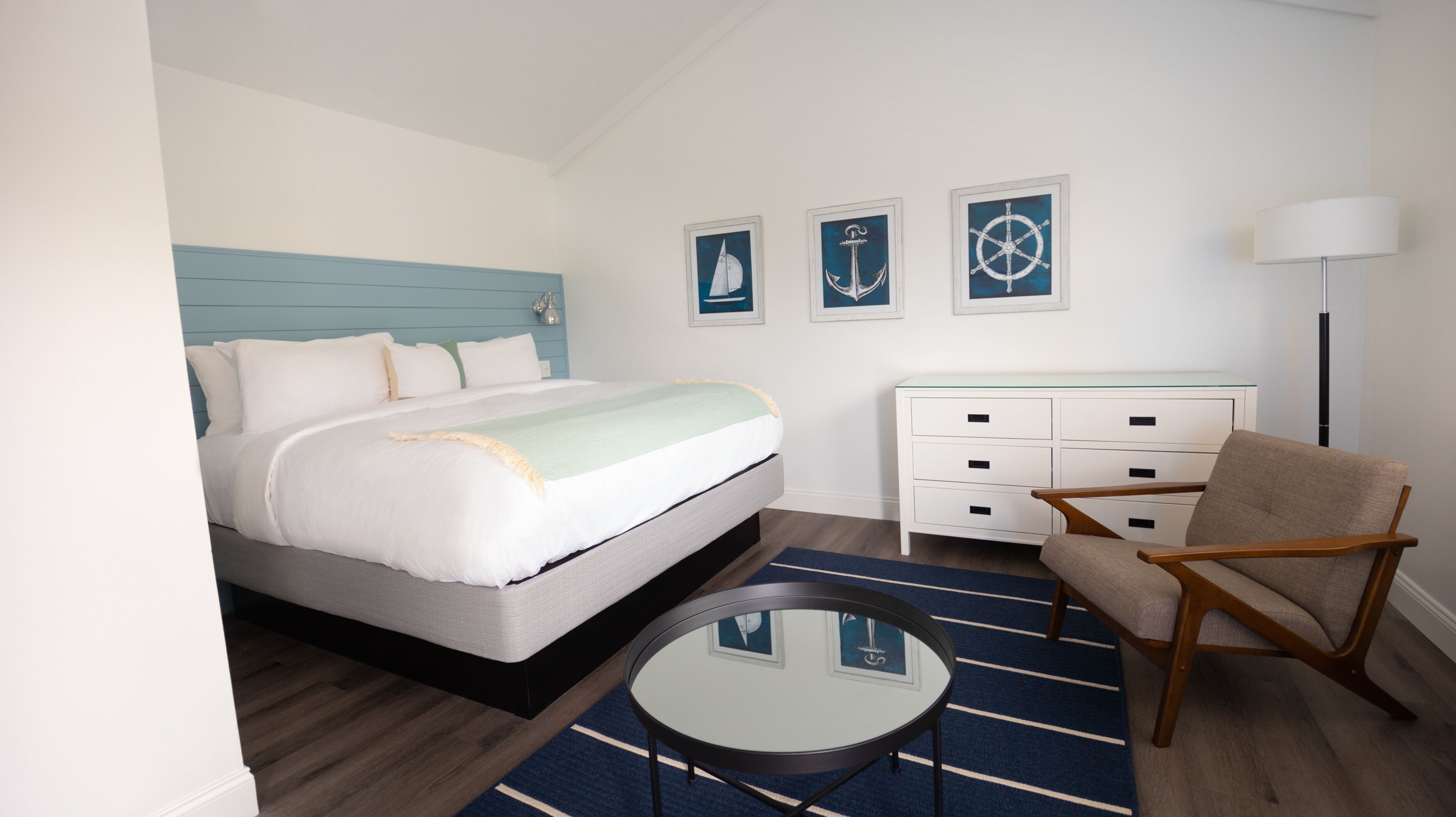 Une chambre d’hôtel bien rangée, avec un lit, une chaise, une petite table et une commode. Trois tableaux encadrés ornent le mur.