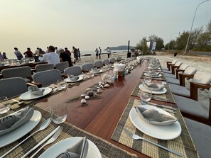 Une longue table à manger, dressée pour un repas sur la plage, avec des chaises et des assiettes disposées en rangées.