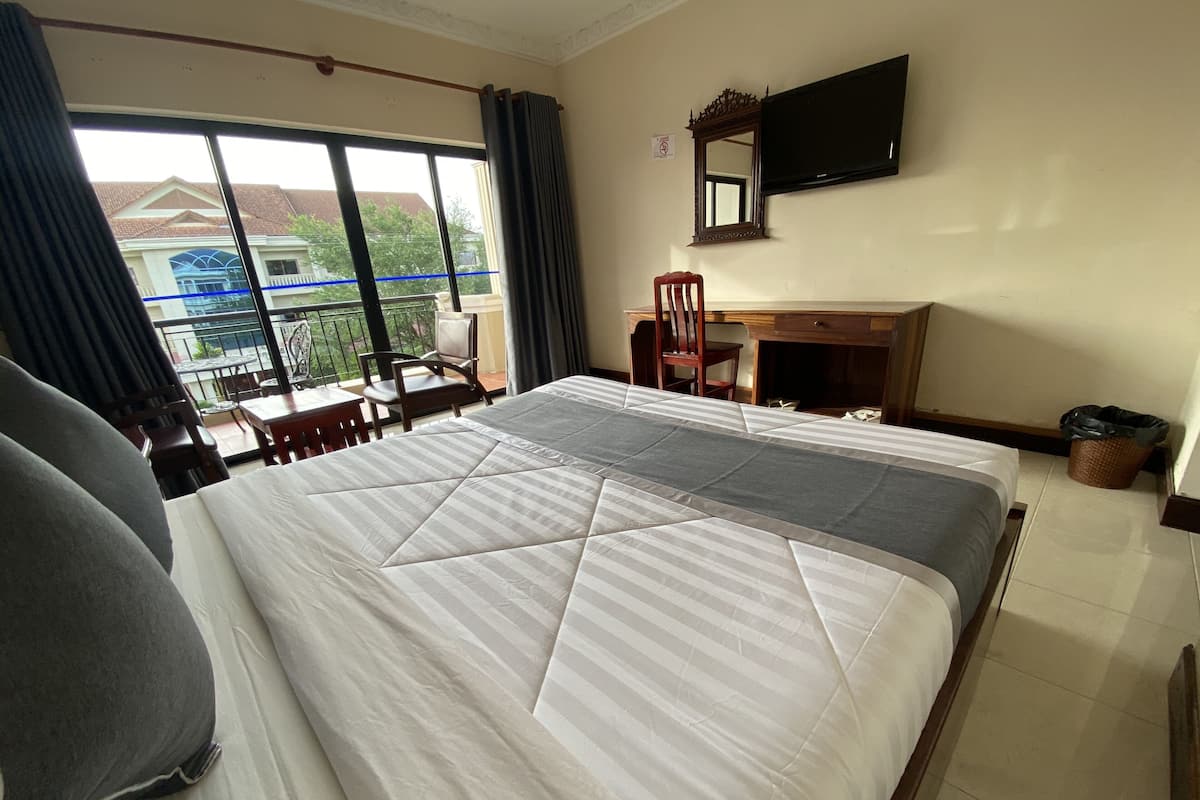 A hotel room with a large bed, a desk, a chair, a TV, and a balcony with a view of buildings.