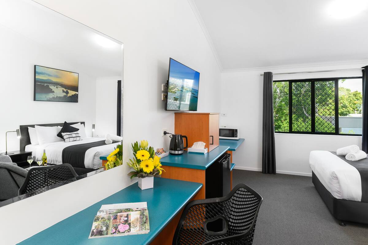 A hotel room with a bed, a desk with a computer, a coffee maker, a TV, and a window with a view of trees.