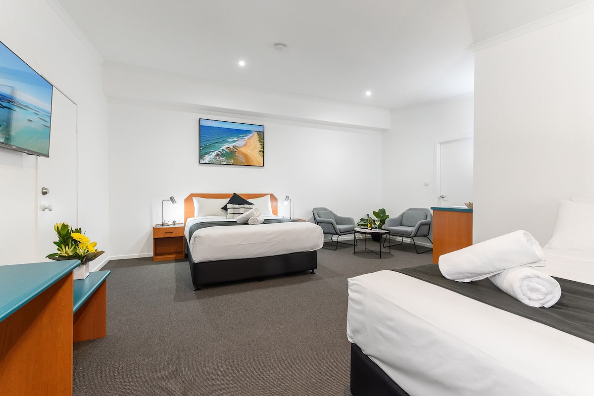 A hotel room with a bed, a desk, a chair, a small table, and a painting on the wall.