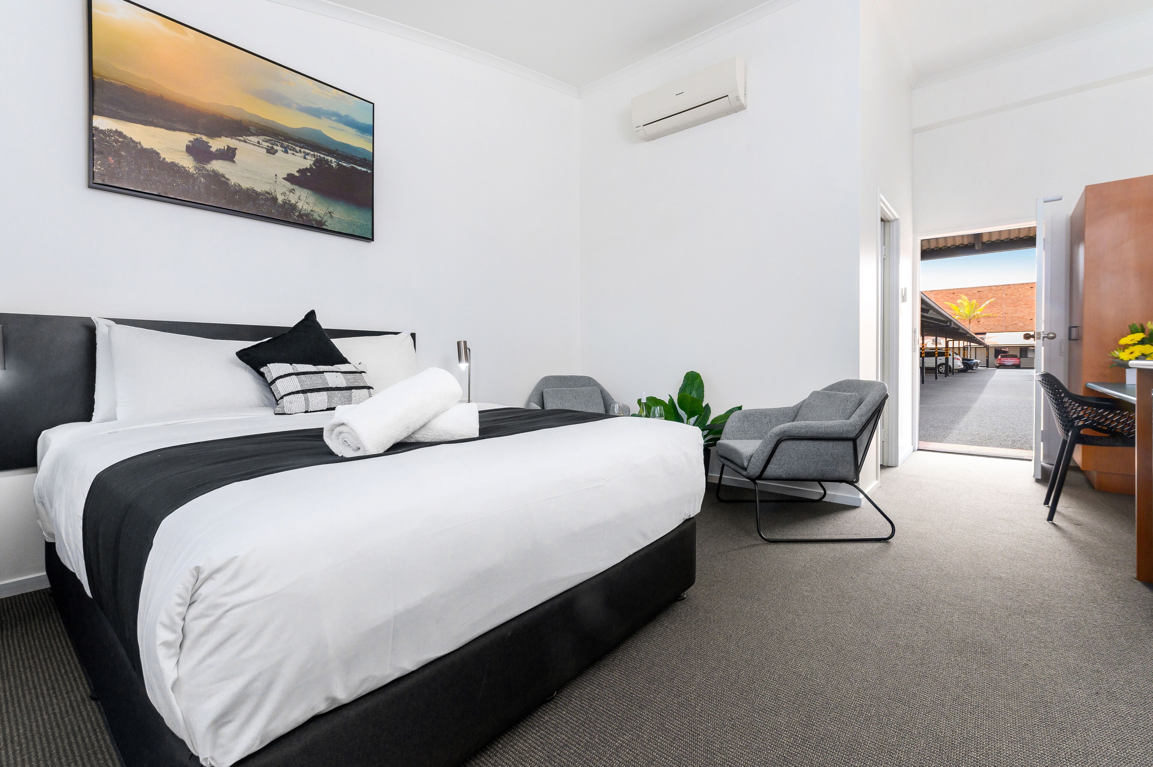 Ein Hotelzimmer mit einem großen Bett, einem Schreibtisch mit Stuhl, einer Zimmerpflanze und einem Bild an der Wand.