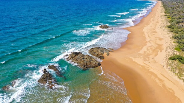 Vista aérea de una playa con aguas azul turquesa, orilla arenosa y afloramientos rocosos.