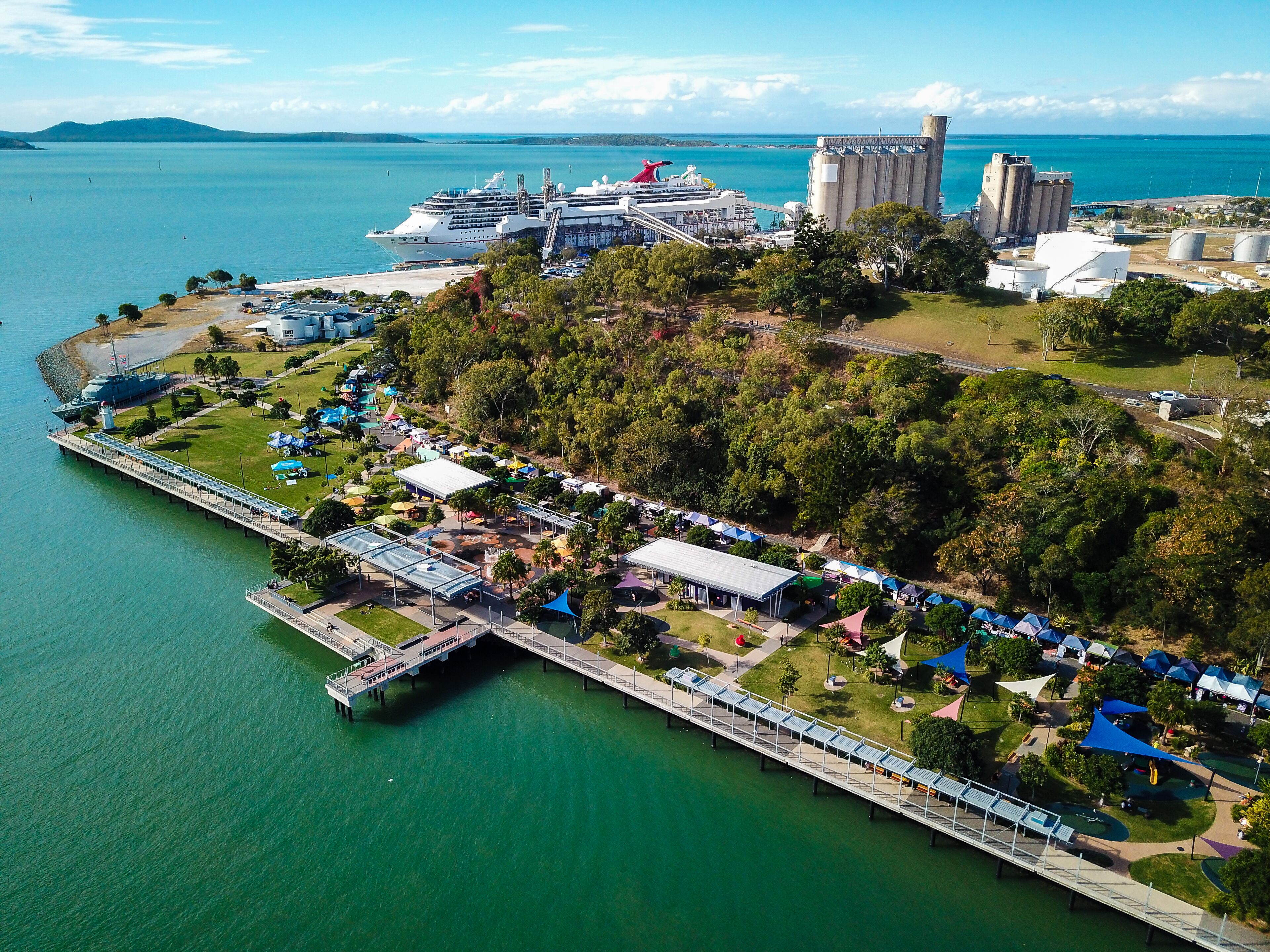 An aerial view of a coastal area with a pier, a cruise ship docked nearby, and a large industrial facility in the background.