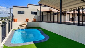 Una piscina con un elemento decorativo de piedra, rodeada de césped artificial y una valla metálica.