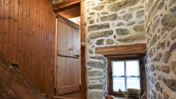 Uma porta de madeira que dá para um quarto com paredes de pedra, uma pequena janela e um banco de madeira.