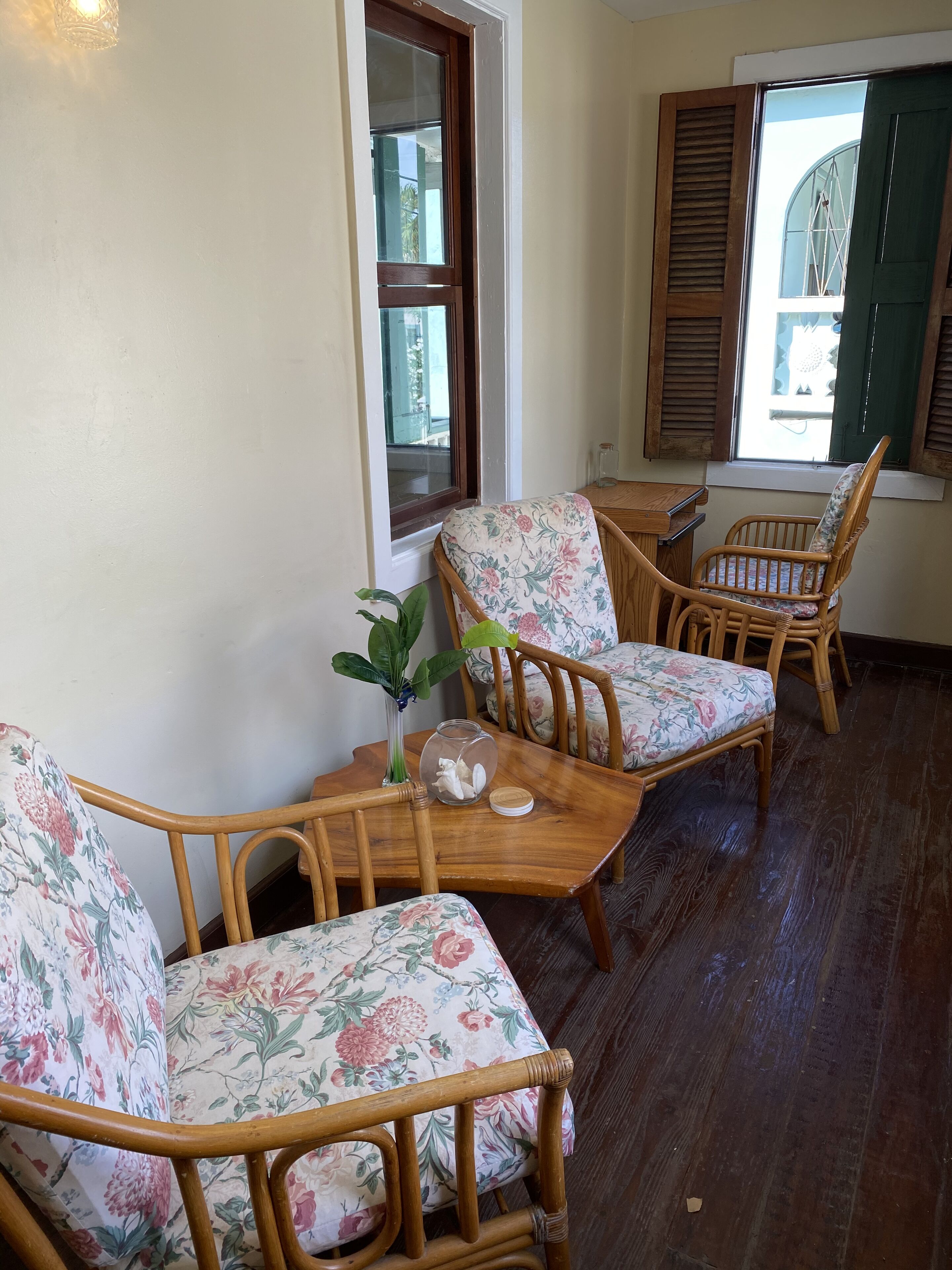 Ein gemütliches Zimmer mit Holzmöbeln, darunter ein kleiner Tisch und zwei Stühle mit floralem Bezug. Auf dem Tisch steht eine Vase mit einer Pflanze, und ein Fenster mit Fensterläden ist sichtbar.
