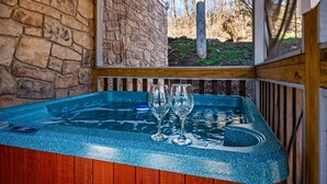 A hot tub with a stone wall and wooden deck, surrounded by a natural landscape. - 3BR Cabin: Hot Tub, Arcade & Game Room, Near Strip (Pigeon Forge)