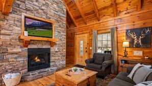 A cozy log cabin interior with a stone fireplace, a television mounted on the wall, a comfortable sofa, and a wooden coffee table. - 3BR Cabin: Hot Tub, Arcade & Game Room, Near Strip (Pigeon Forge)