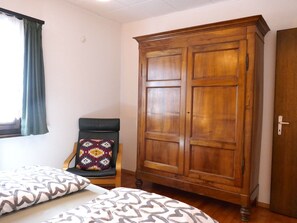 A bedroom with a wooden wardrobe, a chair with a patterned cushion, and two beds with patterned bedding. - Chalet Al Portic by Interhome (Olivone)