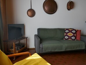 A living room with a television on a wooden stand, a green sofa with a patterned pillow, and two wall-mounted lamps. - Chalet Al Portic by Interhome (Olivone)