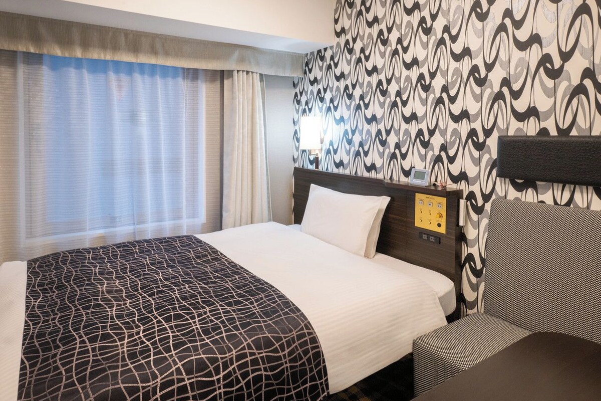 A hotel room with a bed, a bedside table, a control panel, and patterned wallpaper.