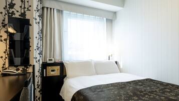 A hotel room with a bed, a desk with a lamp, a clock, and a window with curtains.