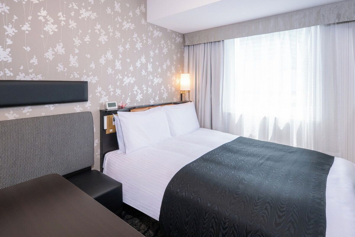 A hotel room with a bed, bedside table, lamp, and a view of the city through the window.