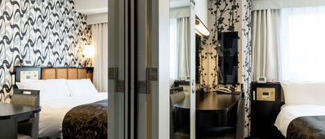 Habitación de hotel con dos camas, un escritorio, una silla, un espejo, baño con lavamanos e inodoro, y un ventanal con cortinas.