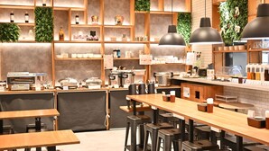Ein modernes Café-Interieur mit Holztischen und schwarzen Barhocker, Regalen mit verschiedenen Artikeln sowie grünen Wanddekorationen.