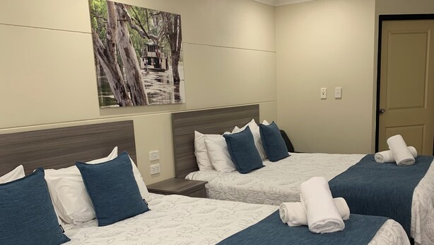 A hotel room with three beds, each with a blue bedspread and white pillows. There is a painting on the wall above the beds, and a door to the right. - Balranald Club Motel (Balranald)