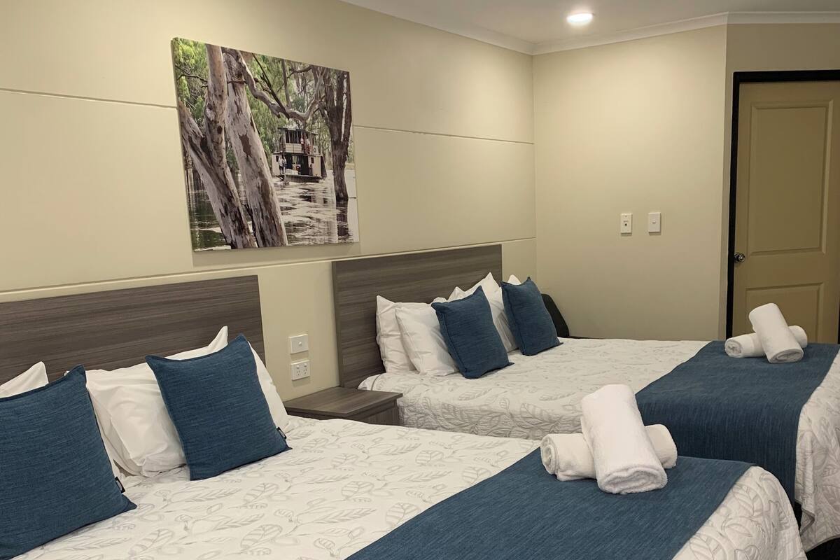 Habitación de hotel con tres camas, cada una con cubrecama azul y almohadas blancas. Hay un cuadro en la pared sobre las camas, y una puerta a la derecha.