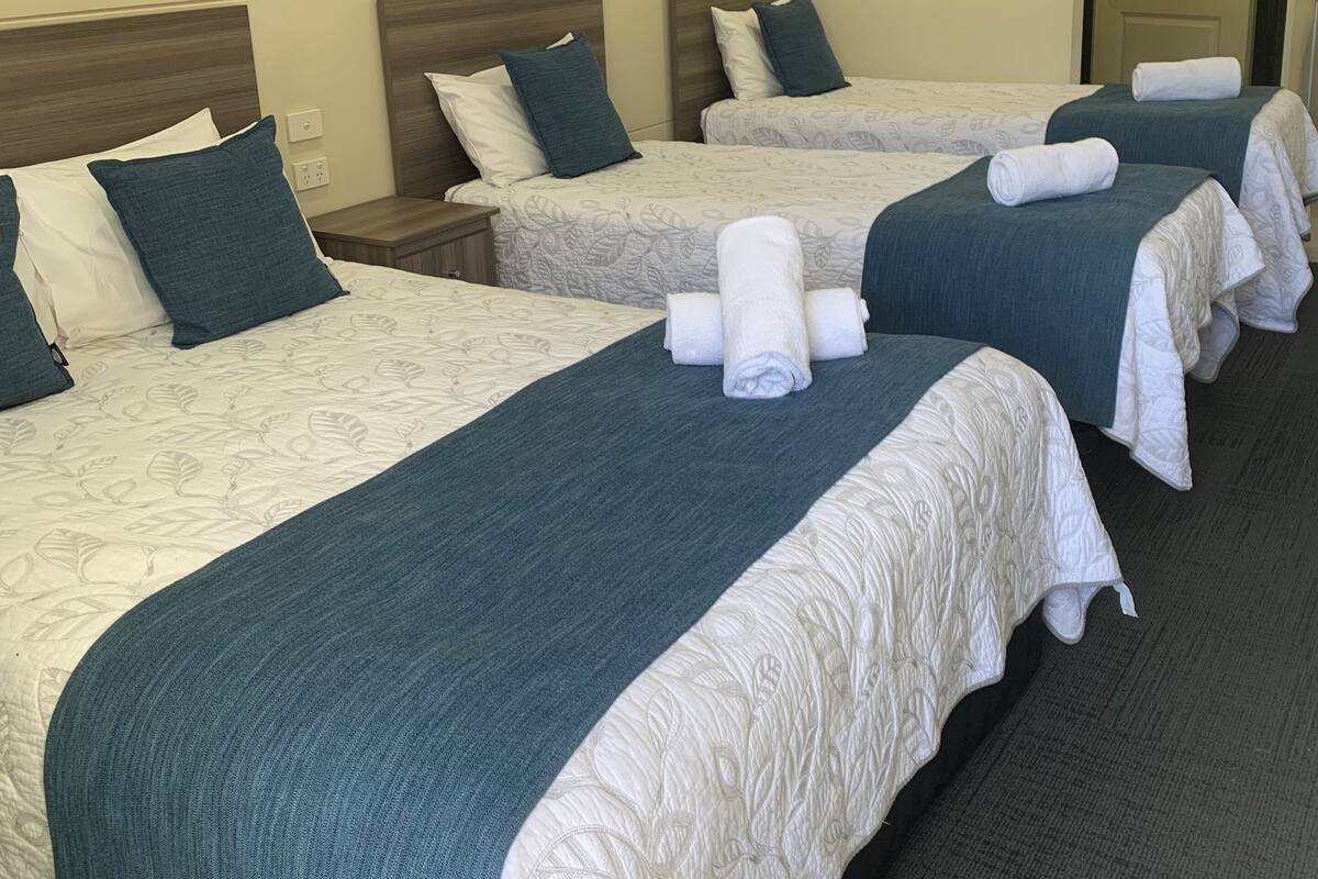 Habitación de hotel con cuatro camas, cada una con una manta azul y almohadas blancas. Se observa una puerta y una mesita con una lámpara.