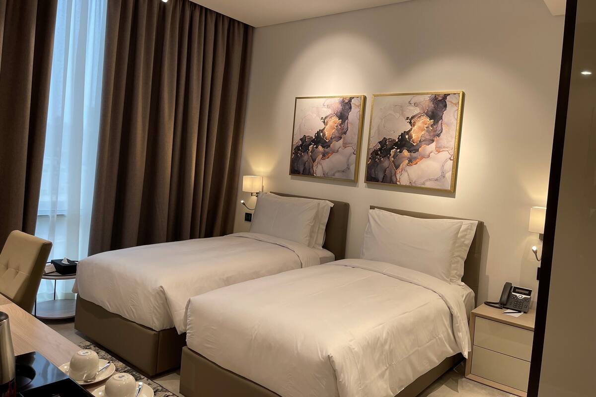 A hotel room with two beds, a desk with a chair, a bedside table with a lamp, and artwork on the wall.